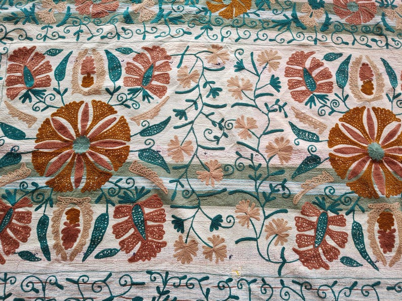 May include: Close-up of a cream-colored textile with intricate embroidery. The design features large, stylized flowers in shades of orange and brown, with teal accents. Delicate vines and smaller flowers fill the spaces, creating a detailed pattern.