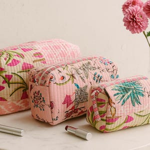 Puede incluir: Tres neceseres acolchados rosas con estampados florales de diferentes tamaños. Los neceseres están sobre una superficie blanca con una vela encendida, dos barras de labios y un jarrón con flores rosas. Cierre de cremallera.