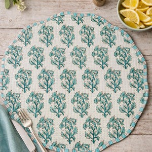 Puede incluir: Mantel individual redondo y acolchado con fondo blanco y un estampado floral turquesa repetido. El borde está rematado con un borde a rayas turquesa y blanco. Tenedor, cuchillo y servilleta azul a la izquierda.