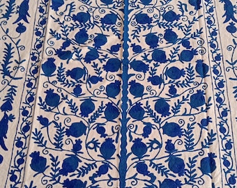 May include: A white textile with a detailed blue embroidered design. The central motif is a stylized tree with fruit, surrounded by a border with floral and bird-like patterns. The embroidery is intricate.