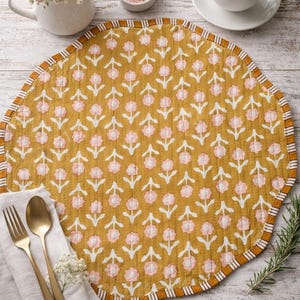 Puede incluir: Mantel individual redondo amarillo mostaza con un estampado floral en rosa y blanco. El mantel individual tiene un borde festoneado con un borde a rayas. Una taza blanca con flores rosas, una taza de café y cubiertos dorados también están sobre la mesa.