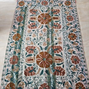 May include: A rectangular, off-white textile with intricate floral embroidery. The design features large, brown and rust-colored flowers with green and teal accents, surrounded by vines and smaller blossoms. The edges are finished with a decorative border.