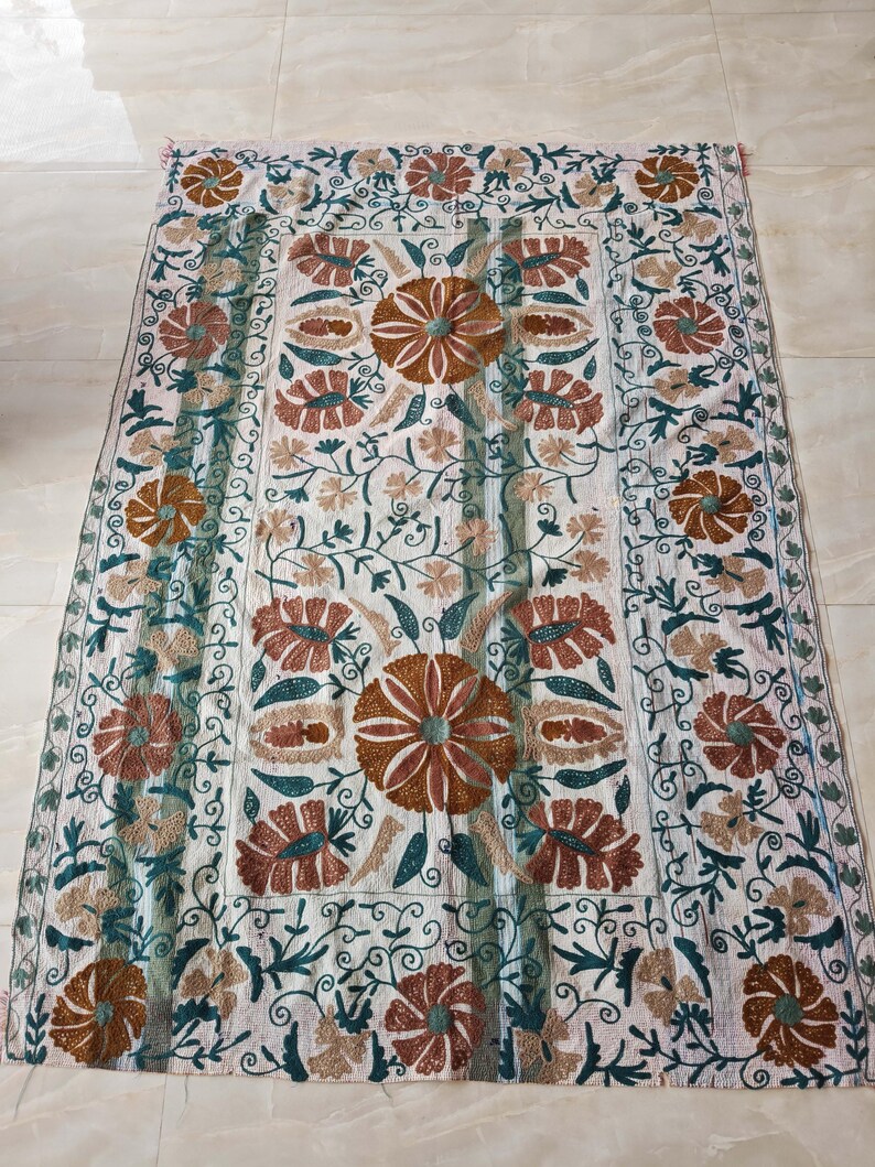 May include: A rectangular, off-white textile with intricate floral embroidery. The design features large, brown and rust-colored flowers with green and teal accents, surrounded by vines and smaller blossoms. The edges are finished with a decorative border.