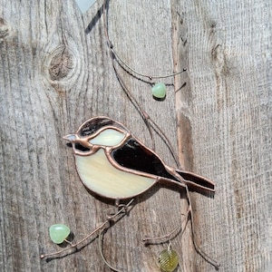 May include: Stained glass bird suncatcher with a black, white, and cream-colored body, perched on a copper wire branch with green glass accents. The bird is hanging from a wooden surface.