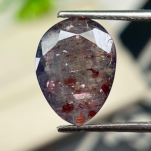 May include: A pear-shaped faceted gemstone with a translucent purplish-grey base colour, featuring distinct internal red and copper-coloured metallic inclusions scattered throughout the stone, held by a pair of silver-toned metal tweezers.