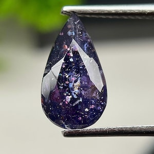 May include: A pear-shaped, faceted gemstone with a deep purple base colour, featuring internal inclusions that create a sparkling, multi-coloured effect with flecks of blue, pink, and silver throughout the translucent, polished stone surface.