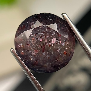 May include: A round, faceted gemstone with a deep purple and dark grey base colour, featuring numerous internal red and metallic inclusions that create a sparkling, glitter-like effect throughout the stone's translucent, polished surface.