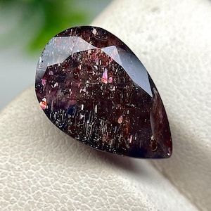 May include: A pear-shaped faceted gemstone with a deep dark red and purple base colour. The stone features internal metallic inclusions that create shimmering flashes of orange, pink, and silver light throughout the dark, translucent body.