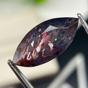 May include: A marquise-cut gemstone with a deep purple and translucent base, featuring internal inclusions of dark red, metallic copper, and iridescent flecks that catch the light. The stone is held by metal tweezers, showcasing its faceted surface.