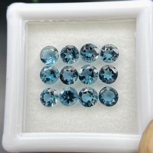 May include: Twelve round, faceted, blue gemstones are presented in a white plastic case. The gems are a deep teal colour and appear to be evenly cut. The case is set against a white background.