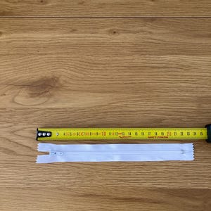 Peut inclure: Une fermeture éclair blanche en nylon mesurant environ 24 cm de long, placée directement sous un mètre ruban jaune indiquant l'échelle en centimètres, avec une petite tirette en métal à l'extrémité supérieure de la fermeture.