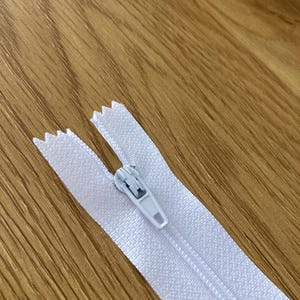 Peut inclure: Une fermeture éclair blanche dotée d'un curseur et d'une tirette en plastique blanc assortis. Le ruban de la fermeture est composé d'un tissu tissé et les dents sont intégrées au ruban, présentant un design standard pour les vêtements.
