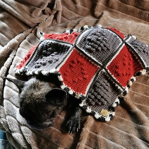 Puede incluir: Un perro bulldog francés marrón y negro está durmiendo debajo de una manta de crochet roja, blanca y gris. La manta tiene un diseño texturizado y un borde blanco.