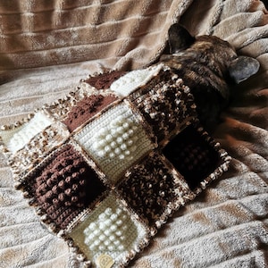Puede incluir: Una manta de crochet marrón y blanca con un patrón texturizado. La manta está hecha de cuadrados con diferentes texturas y colores. La manta está sobre una superficie peluda marrón.