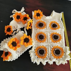 Könnte beinhalten: Ein gehäkeltes Baby-Outfit mit Sonnenblumen-Motiv. Das Outfit besteht aus einer Mütze, Stiefeln und einer Weste. Die Weste ist aus weißem Garn mit braunen und gelben Sonnenblumen-Akzenten gefertigt. Die Mütze und die Stiefel sind weiß mit gelben Sonnenblumen-Akzenten.