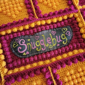 May include: A close-up of a crocheted blanket with a yellow and purple pattern. The blanket has a label that says "Snugglebug" with hearts around the text.