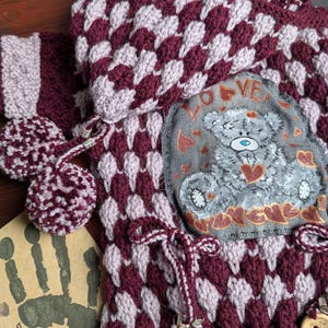 Può includere: Un cardigan con cappuccio in maglia color bordeaux e rosa con un'applicazione a forma di orsacchiotto di peluche con la scritta "LOVE". Il cardigan ha una chiusura con coulisse e bottoni in legno.