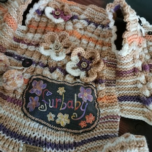 Può includere: Un maglione di lana per neonato a righe beige, arancio e viola con una toppa di feltro marrone che dice "Surbaby" in lettere colorate. Il maglione ha fiori di uncinetto bianchi e un'etichetta che dice "fatto a mano da Kiholino".