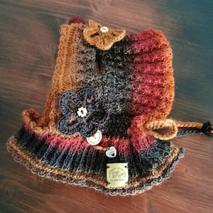 Peut inclure: Un chapeau crocheté avec un dégradé de couleurs brun, noir et rouge. Le chapeau est orné d'une fleur crocheté et d'une étiquette qui indique "Handmade with Love by Klobimo".