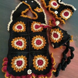 May include: A crocheted set in black, burgundy, and gold featuring a vest, a hat, and a neck warmer. The vest has a scalloped edge and is made of granny squares. The hat has a long, crocheted cord.