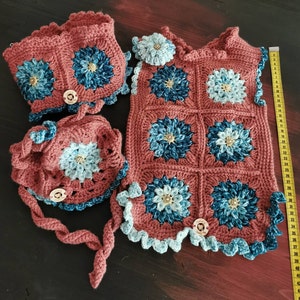Può includere: Un set da neonato all'uncinetto in tonalità marrone, blu e bianco. Il set comprende un cappello, un giubbotto e un scaldacollo. Il giubbotto ha un motivo quadrato con un design floreale.