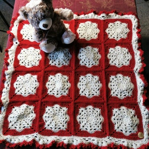 Può includere: Una copertina per neonati in crochet rosso e bianco con un bordo a festoni. La copertina è fatta di quadrati con un disegno di fiore bianco al centro di ogni quadrato. Un orsacchiotto di peluche marrone è seduto sopra la copertina.