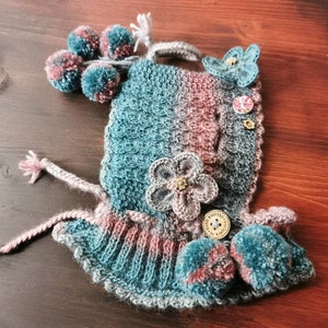 May include: A teal and pink knitted hat with a crocheted flower and pom poms. The hat has a button with the text "Hand Knitted" on it.