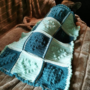 May include: A crocheted blanket with a blue and green checkered pattern. The blanket is made with a textured stitch and has a scalloped edge.