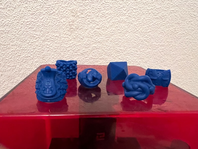 May include: Six blue 3D printed rings on a red surface. The rings are different shapes and sizes.