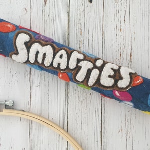 May include: A felted blue Smarties candy bar with white lettering and colourful candy accents. The word "Smarties" is prominently displayed. The candy bar is lying on a white wooden surface, with a wooden embroidery hoop in the lower left corner.