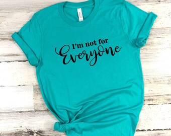 May include: Teal t-shirt with the phrase "I'm not for Everyone" in black script. The shirt is made of soft material and has rolled-up sleeves. The bottom of the shirt is tied in a knot.