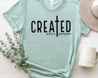 Puede incluir: Una camiseta verde claro con una cruz negra y el texto "CREATED with a purpose".