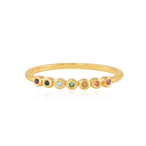 Puede incluir: Un anillo de banda de oro con siete pequeñas piedras redondas de diferentes colores. Las piedras están dispuestas en una línea a través de la banda.