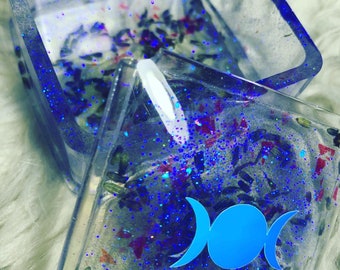 Può includere: Due scatole quadrate trasparenti riempite di glitter, fiori secchi e un simbolo di tripla luna blu. Le scatole sono impilate l'una sull'altra.