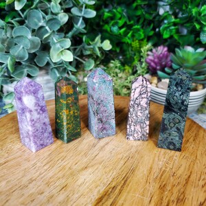May include: Five polished crystal points in various colors and patterns. The points are arranged in a row on a wooden surface. The colors include purple, green, pink, and a mix of green and brown.