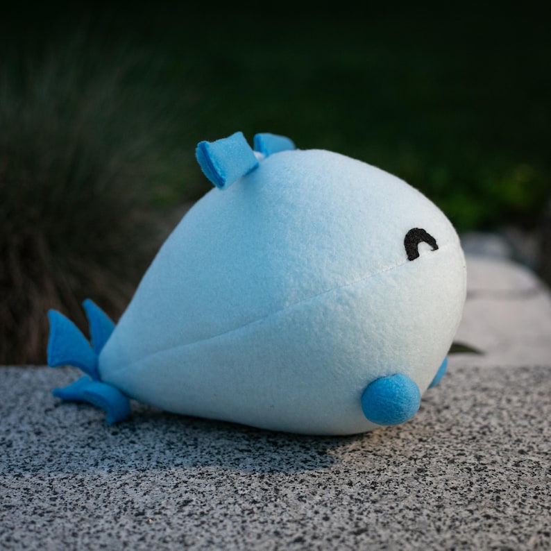 May include: A light blue plush whale toy with a rounded body, small black eye, and blue fins. The whale has a light blue body with darker blue accents on the fins and top of the head. The toy is made of soft fabric.