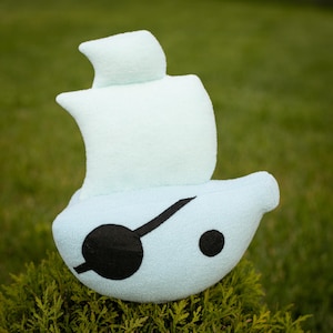 May include: A light blue plush toy sailboat with a black eye patch and a black dot. The sailboat has a white sail and is sitting on green foliage. The toy is made of soft material.