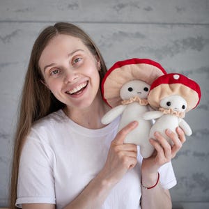 May include: Two mushroom-shaped plush dolls. The dolls have white bodies, small arms, and legs, with red and tan mushroom caps. Each doll has a small, ruffled collar. The dolls are held in front of a light gray background.