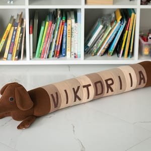 May include: A brown and tan dachshund-shaped plush toy with the name "VIKTORIA" embroidered on its body. The toy is lying on a white tiled floor, with a bookshelf filled with books in the background.