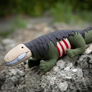 Puede incluir: Un juguete de peluche de dinosaurio con cabeza beige, cuerpo verde y espalda gris deshilachada. El vientre del dinosaurio presenta rayas rojas y blancas. Tiene una sonrisa cosida y está posado sobre una superficie rocosa.