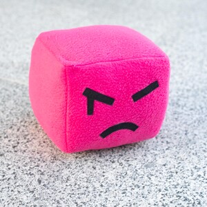 May include: A plush, magenta cube with a black, angry face. The cube has a soft, textured surface and a simple, minimalist design. The face features two angled lines for eyebrows and a curved line for a mouth.