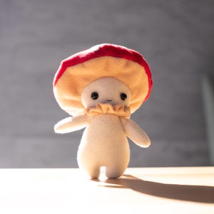 May include: A plush mushroom figure with a red and tan cap, a cream-colored body, and a ruffled collar. The mushroom has small black eyes and outstretched arms, standing on a wooden surface.