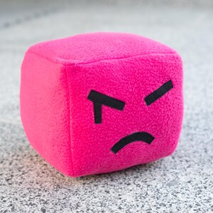 May include: A plush, magenta cube with a stitched, angry face. The cube has black, angular eyebrows and a curved, black mouth, conveying a sense of frustration. The soft, textured material suggests a toy or decorative item.