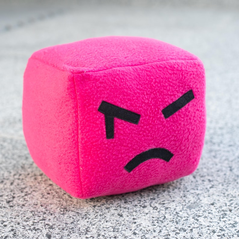 May include: A plush, magenta cube with a stitched, angry face. The cube has black, angular eyebrows and a curved, black mouth, conveying a sense of frustration. The soft, textured material suggests a toy or decorative item.
