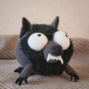May include: A stuffed wolf toy with oversized white eyes, a grey snout, and sharp white teeth. The wolf has black fur and grey paws. The toy is on a textured, light brown surface.