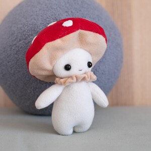May include: A plush mushroom figure with a red cap dotted with white spots. The mushroom has a beige stem and a white body with small arms and legs. It has black eyes and a ruffled beige collar. The background includes a gray pillow and a wooden surface.