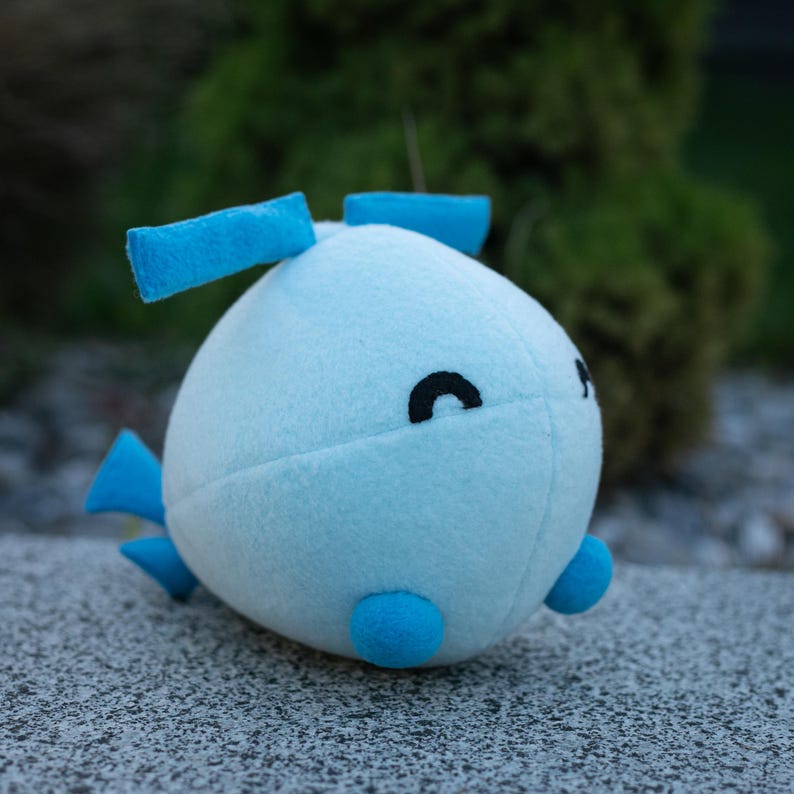 May include: A light blue plush toy with a rounded body, featuring small blue fins and a pair of blue rectangular shapes on top. The toy has a black crescent-shaped eye and a small black dot for a mouth. The toy is sitting on a gray surface.