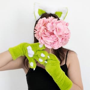 May include: A green plush claw-like gloves with white claws, a green and white cat ear headband, and a pink hydrangea flower. The gloves are a bright green color and have a soft, furry texture. The cat ears are white with green accents.