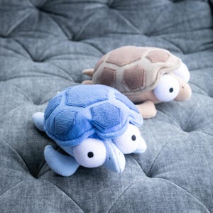 Pode incluir: Dois brinquedos de pelúcia de tartaruga. Um é azul claro com uma carapaça azul mais escura, o outro é bege com uma carapaça marrom. Ambos têm olhos brancos grandes com pupilas pretas. As tartarugas estão em uma superfície cinza acolchoada.