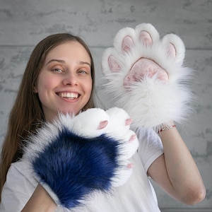 May include: A pair of plush animal paw gloves featuring long faux fur. One glove is solid white with light pink paw pads and claws, while the other glove is dark blue with white fur accents, white paw pads, and light pink claws on each finger.